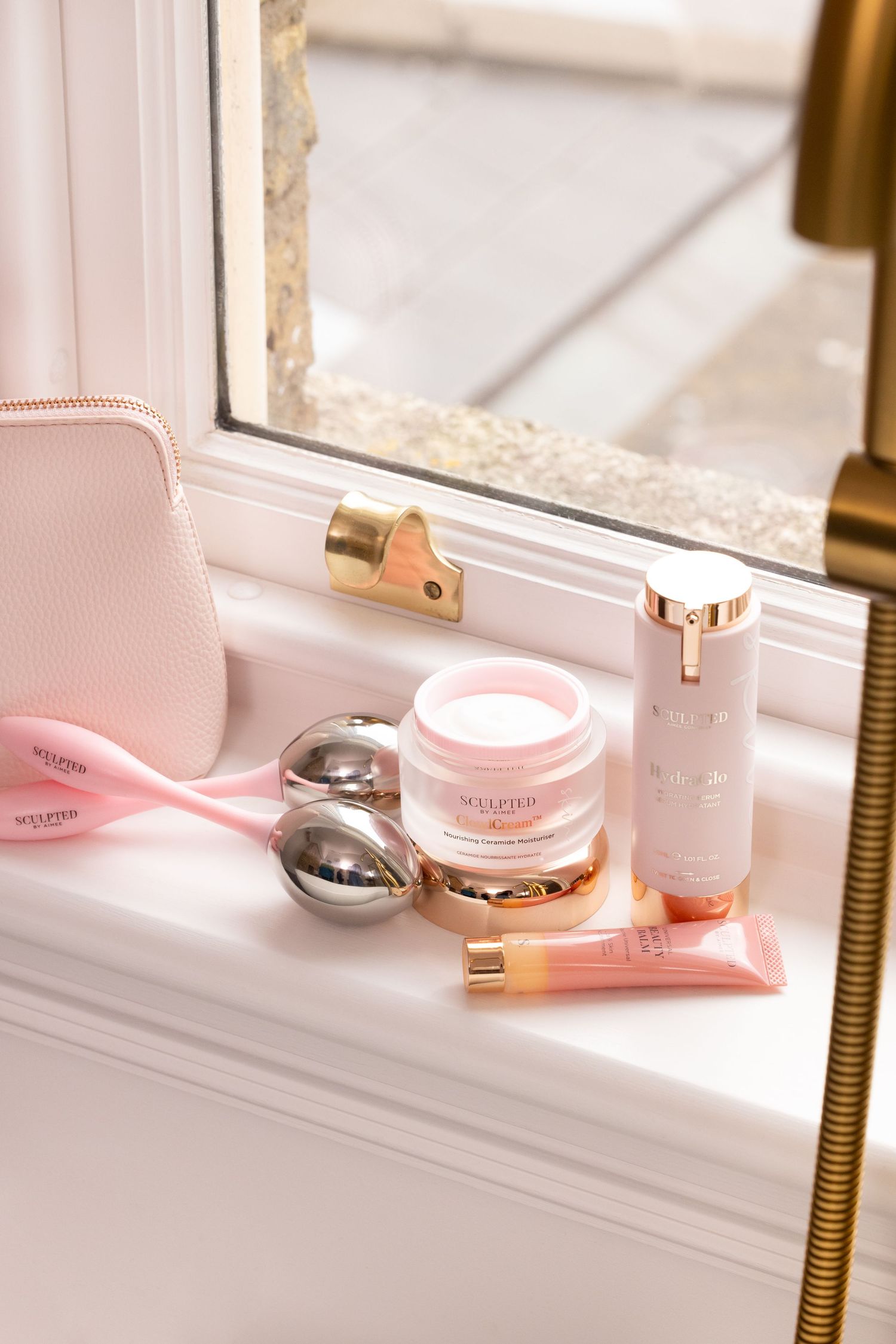 Pink beauty skincare products including Seventh cosmetics bottle and facial tools arranged on white windowsill in natural light.