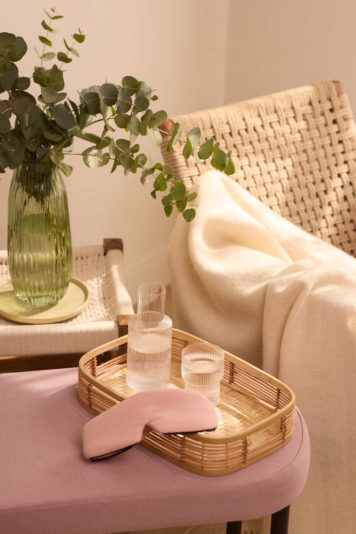Cozy home spa setup with eucalyptus in green vase, woven tray with essentials, and plush towel on pink surface.