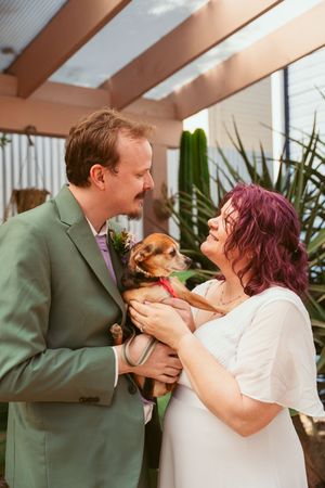 A couple in wedding attire smiles at each other while holding a small brown dog at an indoor ceremony venue.