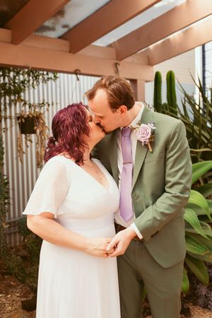 A newlywed couple shares an intimate kiss in a garden setting with greenery and soft lighting.