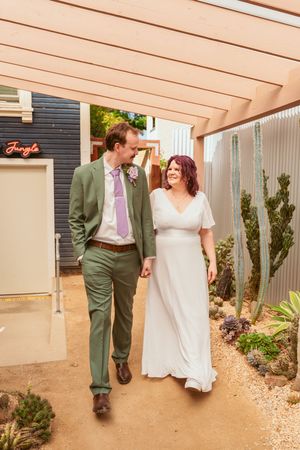 A couple wearing wedding attire walks together on a sandy path surrounded by desert plants and a dark blue wall.