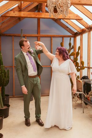 A playful dancing moment between newlyweds in a greenhouse setting with exposed wooden beams and a chandelier.
