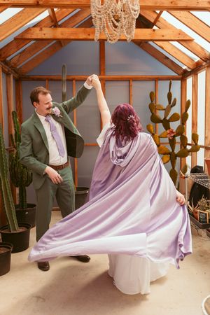 Two people dance together in a greenhouse with exposed wooden beams and desert plants under a crystal chandelier.