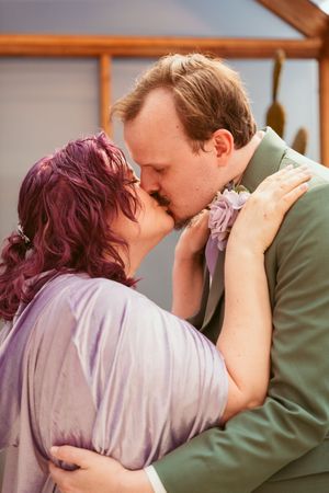 A romantic wedding kiss between a couple, with the bride wearing purple hair and a light dress in a sunlit venue.