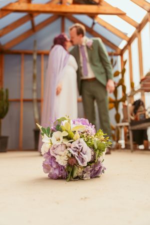 A beautiful wedding bouquet sits in the foreground with purple and white flowers while two people share a moment in a sunlit room.