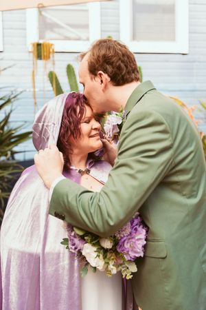 A tender embrace between two newlyweds surrounded by white and purple flowers during an outdoor wedding ceremony.