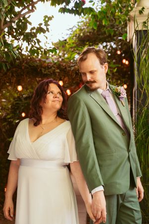 A couple in wedding attire, with the bride in white and groom in sage green, hold hands under garden lights and foliage.