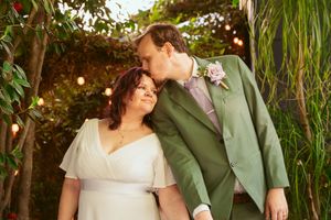 Wedding couple shares an intimate moment in a garden setting, with the groom in a sage green suit kissing bride's head.