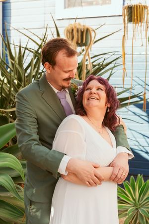 A romantic wedding portrait shows a joyful couple embracing outdoors among tropical plants and decorative elements.