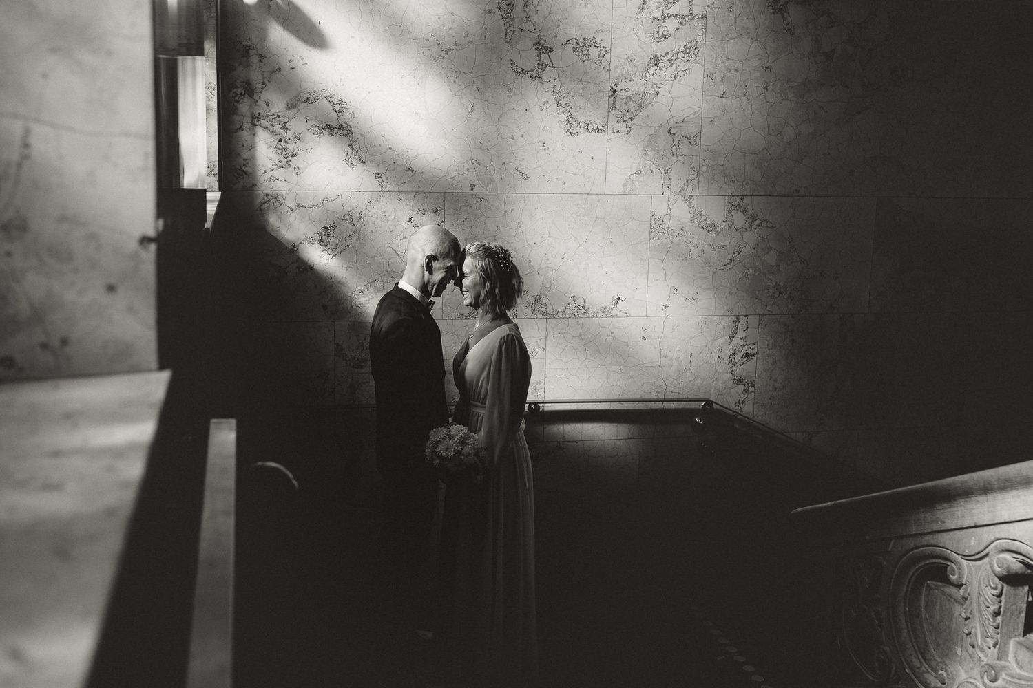 Black and white artistic wedding photo with dramatic lighting and shadows falling on a stone wall backdrop.