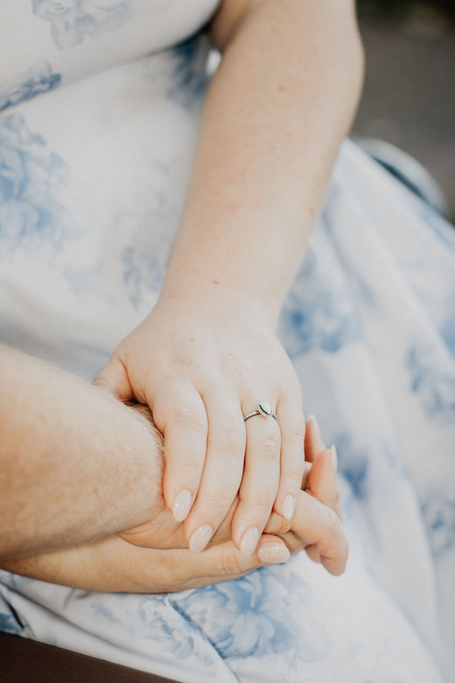 Close-up of two people holding hands, woman wearing a delicate engagement ring over a blue floral dress.