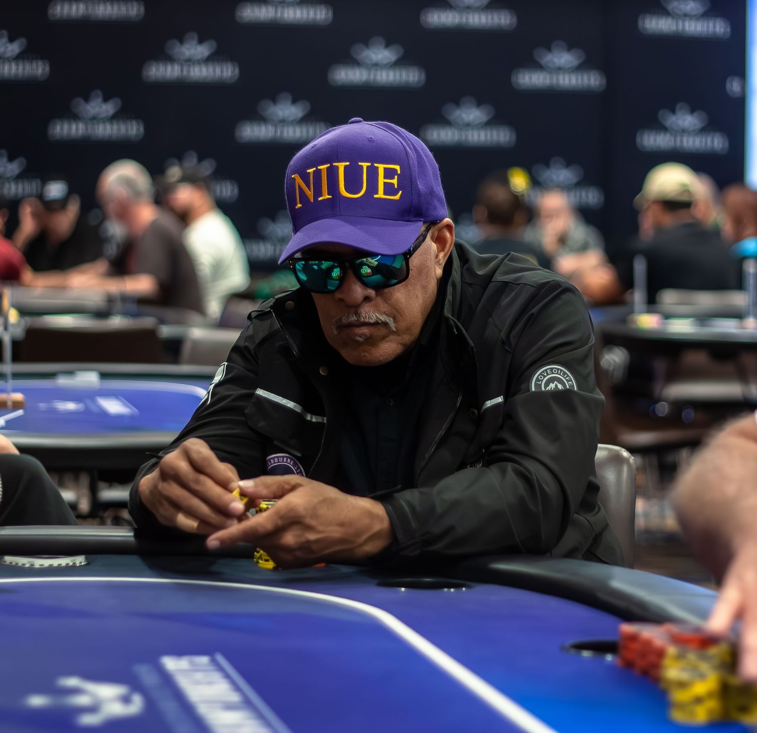 Players sit at a blue poker table wearing sunglasses and dark clothing during a tournament.