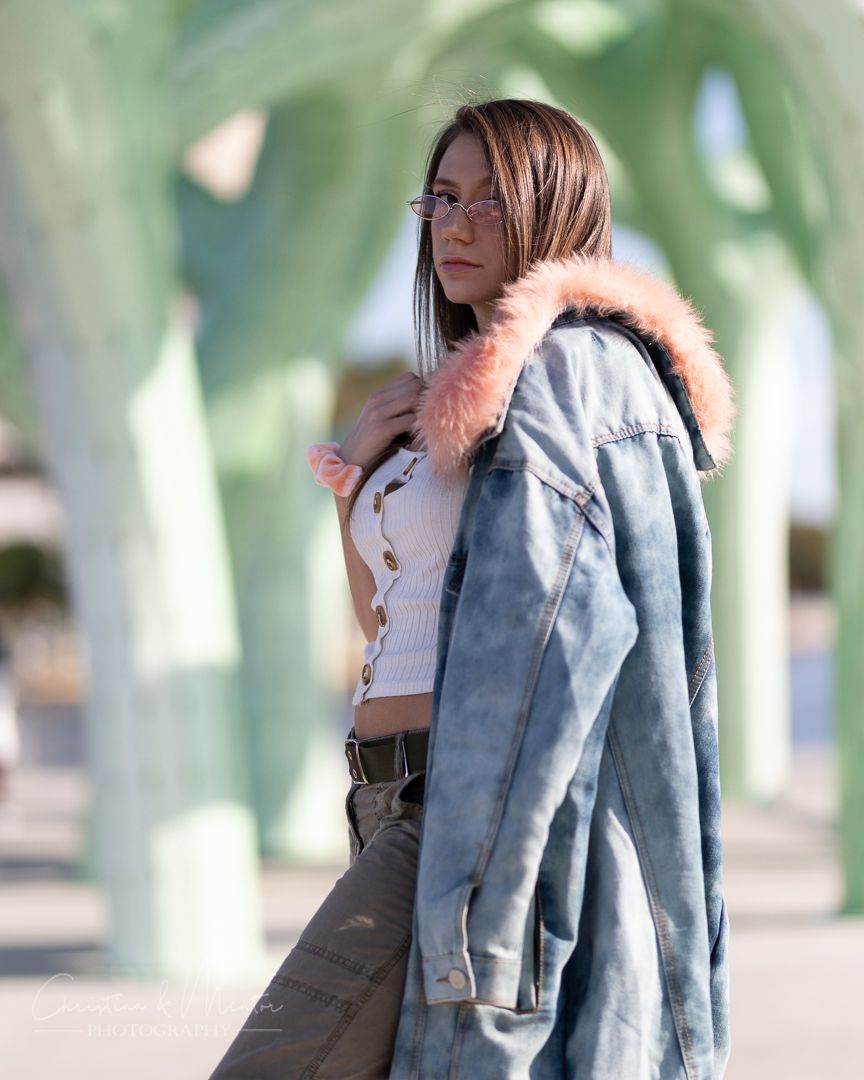 Casual fashion pose in denim jacket with pink fur trim against blurred green foliage.