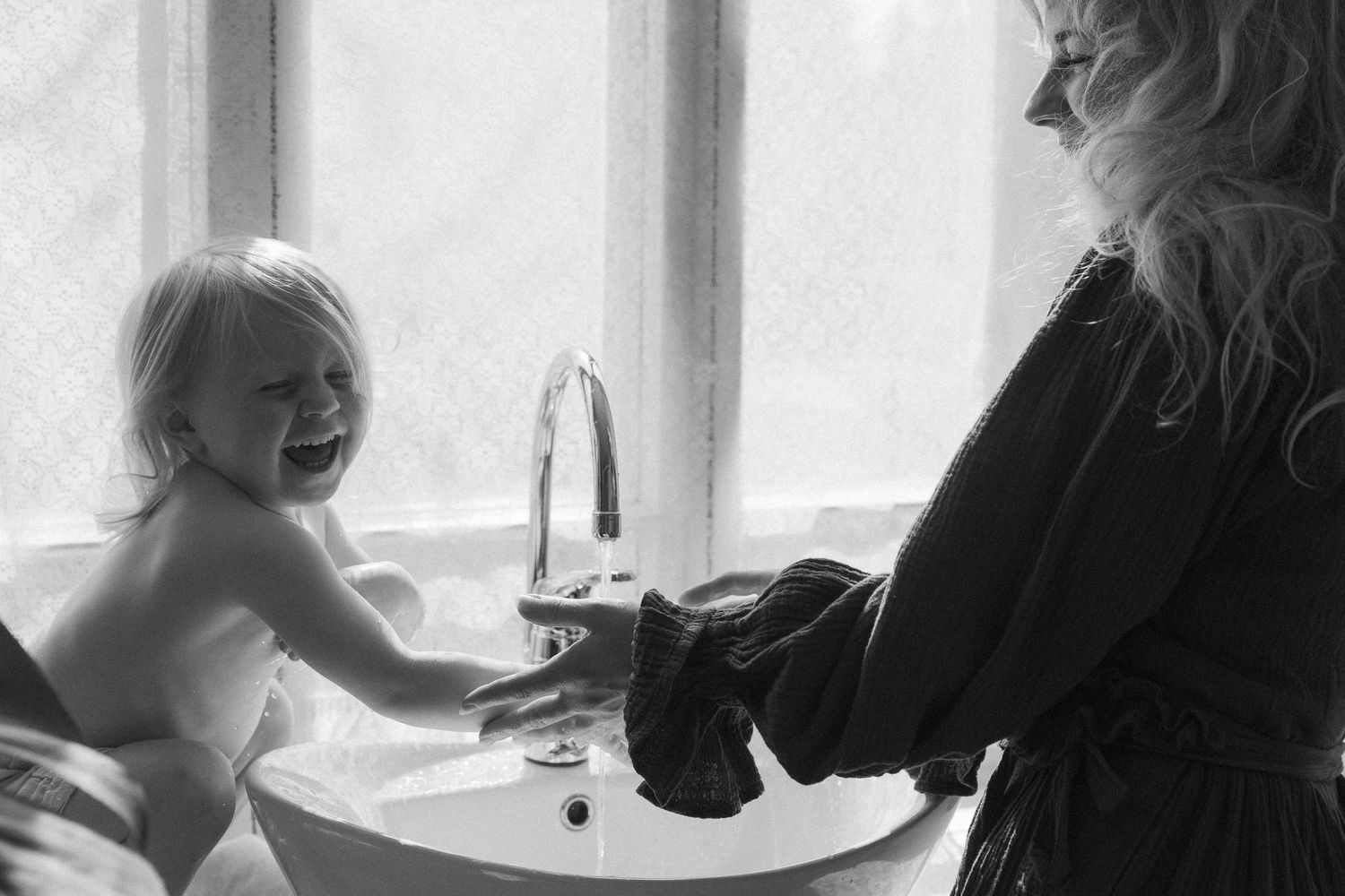 A joyful bathtime moment captured in black and white as a baby laughs while being bathed near a window.