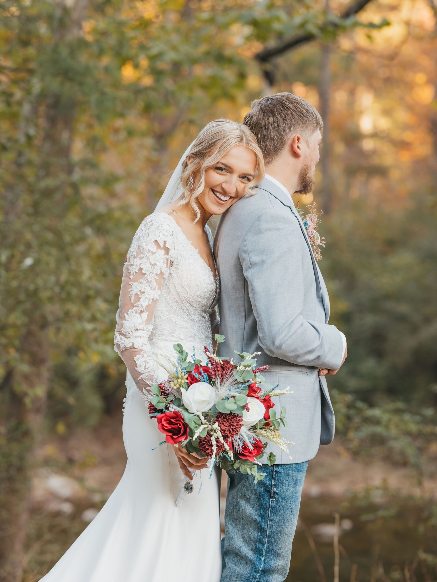 Bride holding red and white bouquet poses with groom in casual wedding attire.