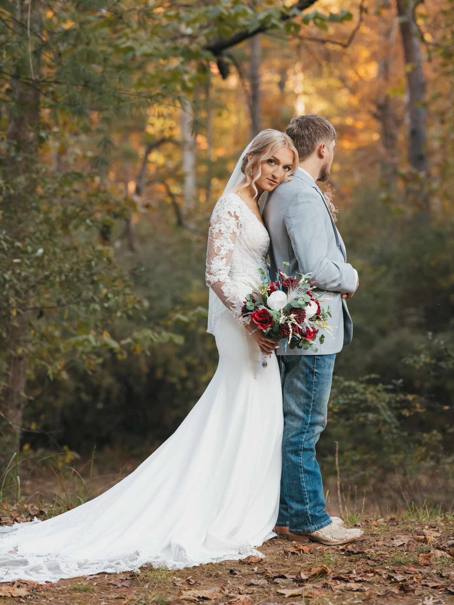 Wedding couple poses with red and white bouquet against autumn forest backdrop.