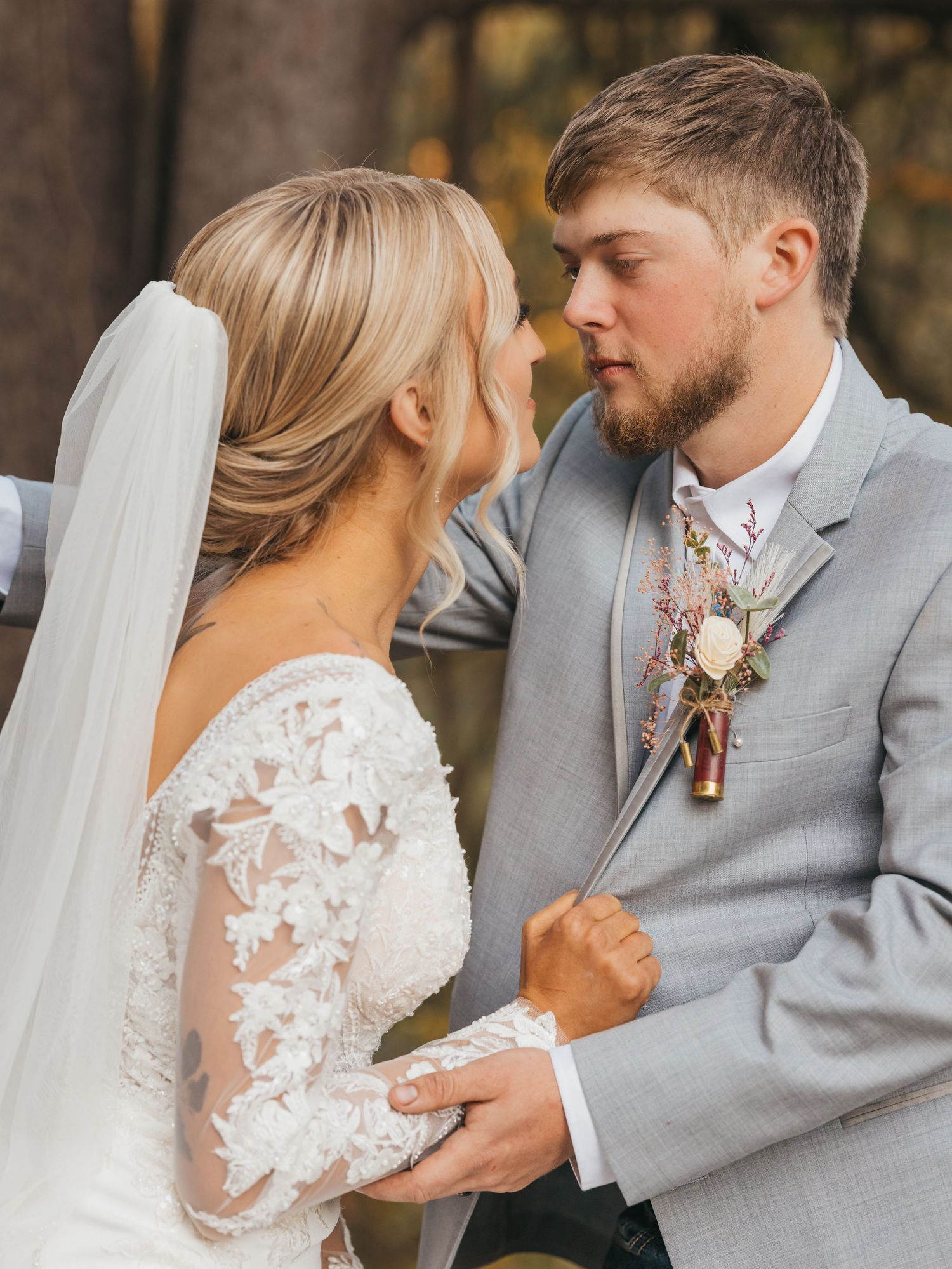 A tender moment between newlyweds with the bride in lace sleeves and groom in grey suit against fall foliage.