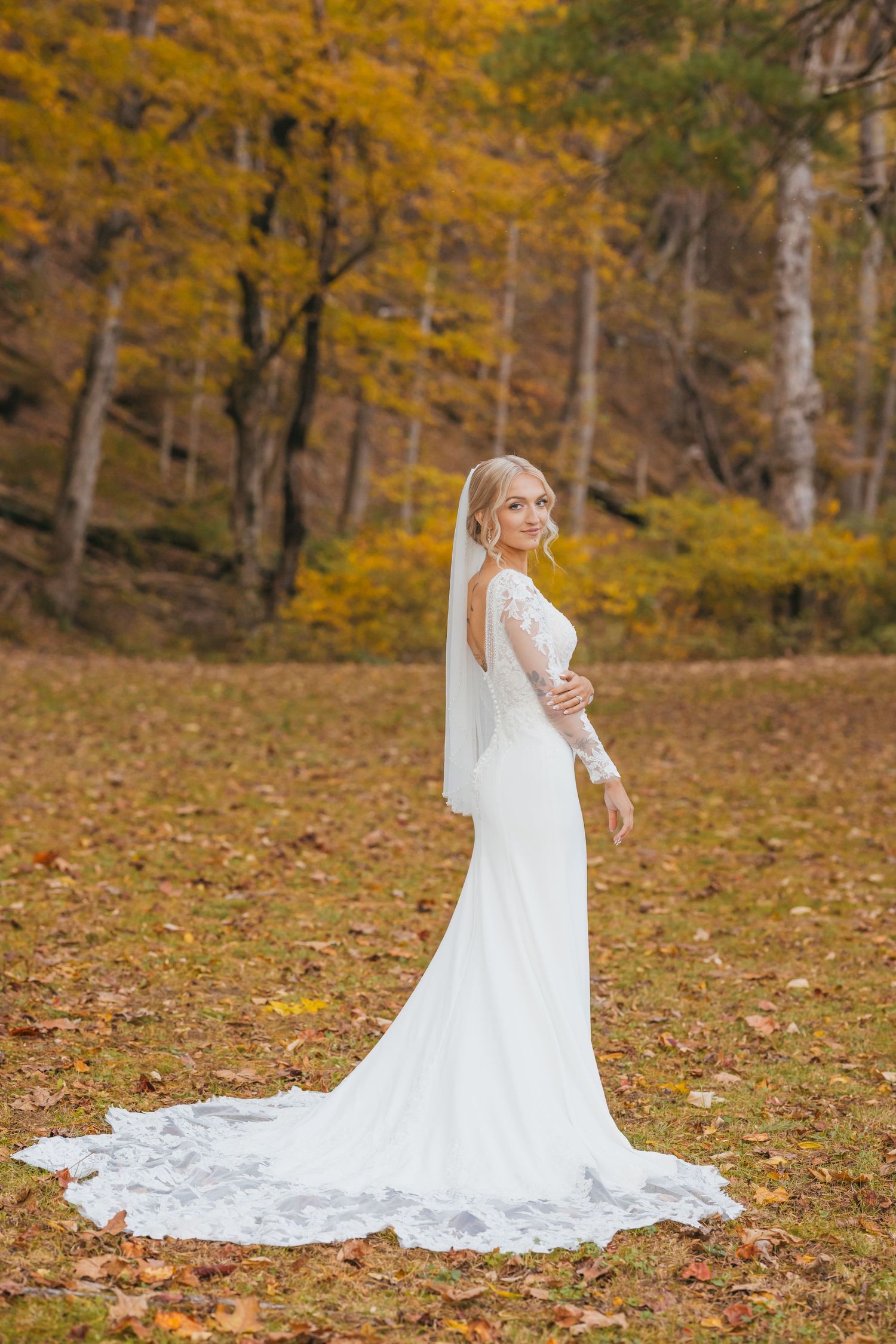 A bride in a fitted lace wedding gown poses in a scenic fall setting with golden leaves.