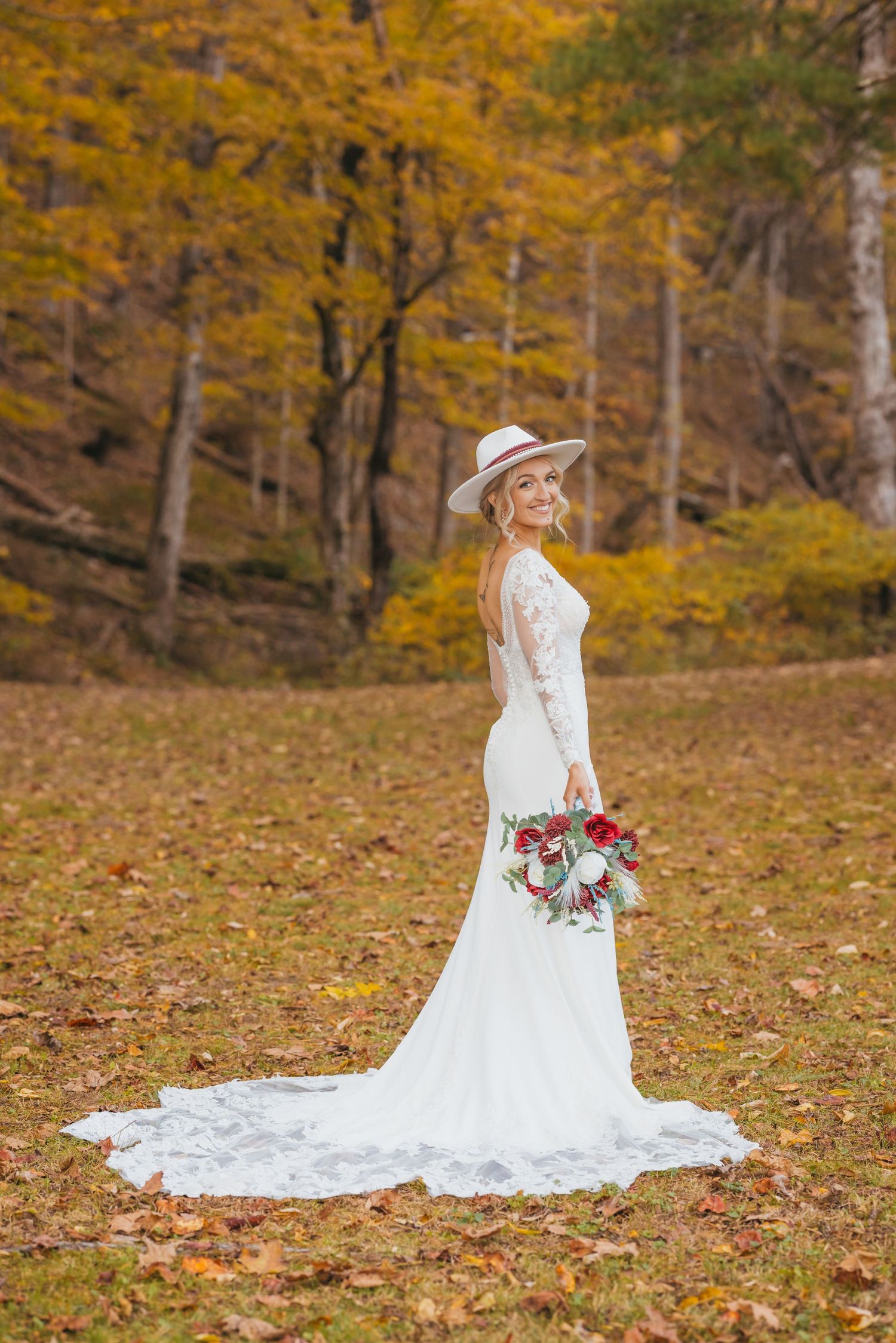 Bride in a flowing white gown and hat poses elegantly among fall foliage.