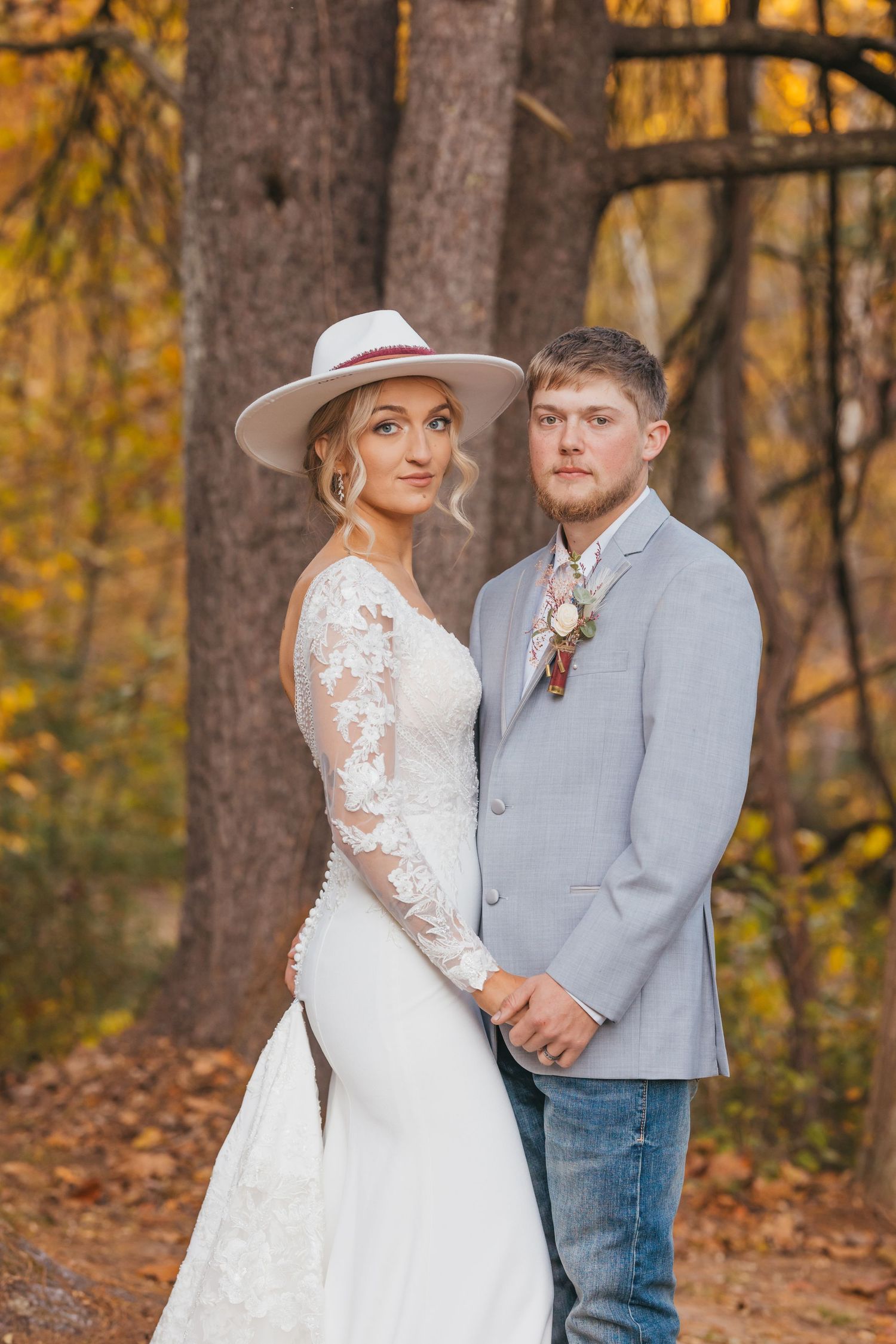 A romantic autumn wedding portrait featuring a bride in lace and groom in light gray against golden fall foliage.