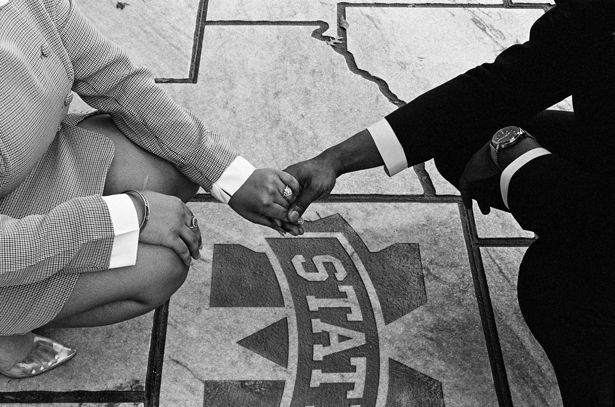 Black and white close-up of two people holding hands over a State University logo on pavement.