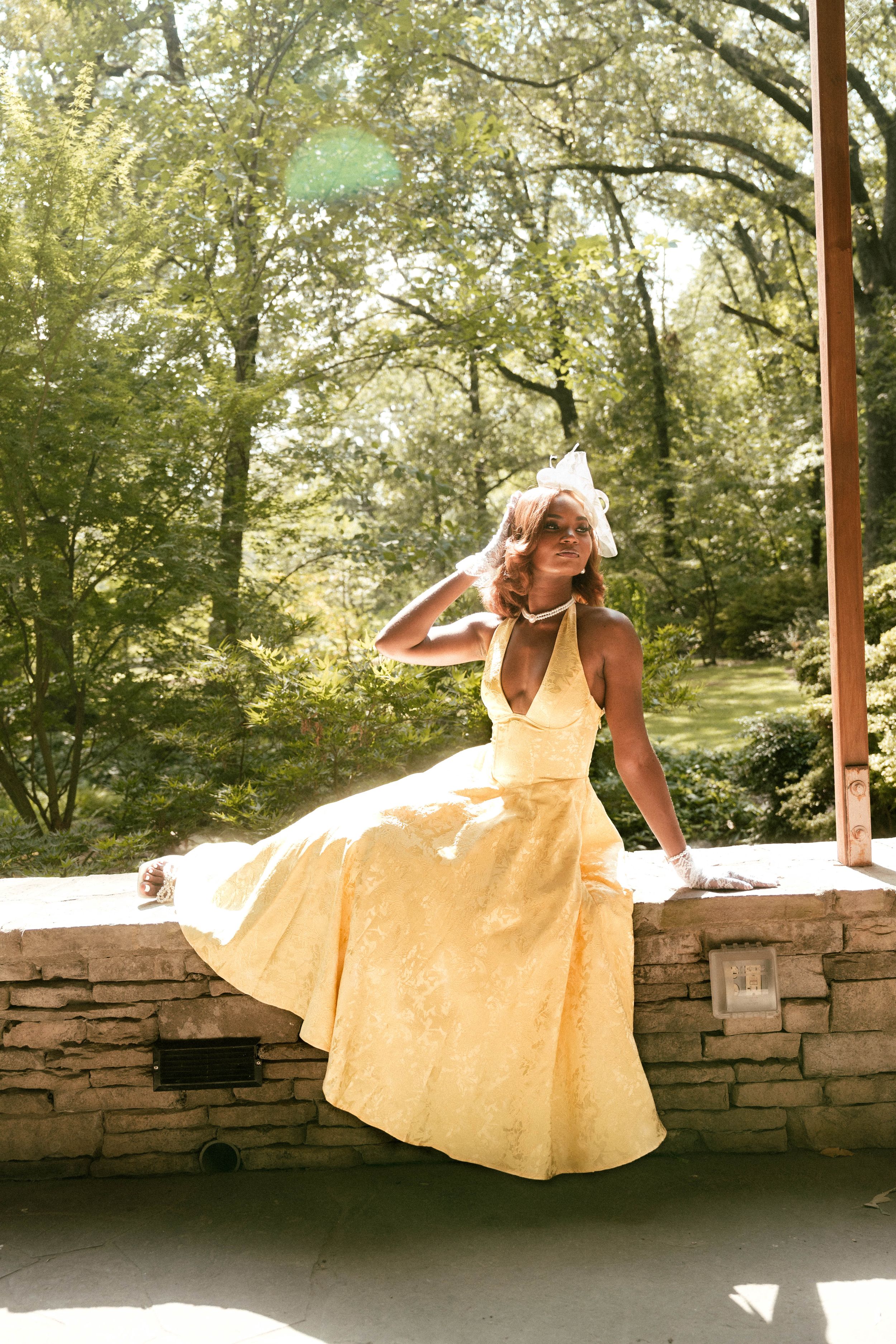 Woman in yellow halter gown sitting on stone wall surrounded by lush green trees.