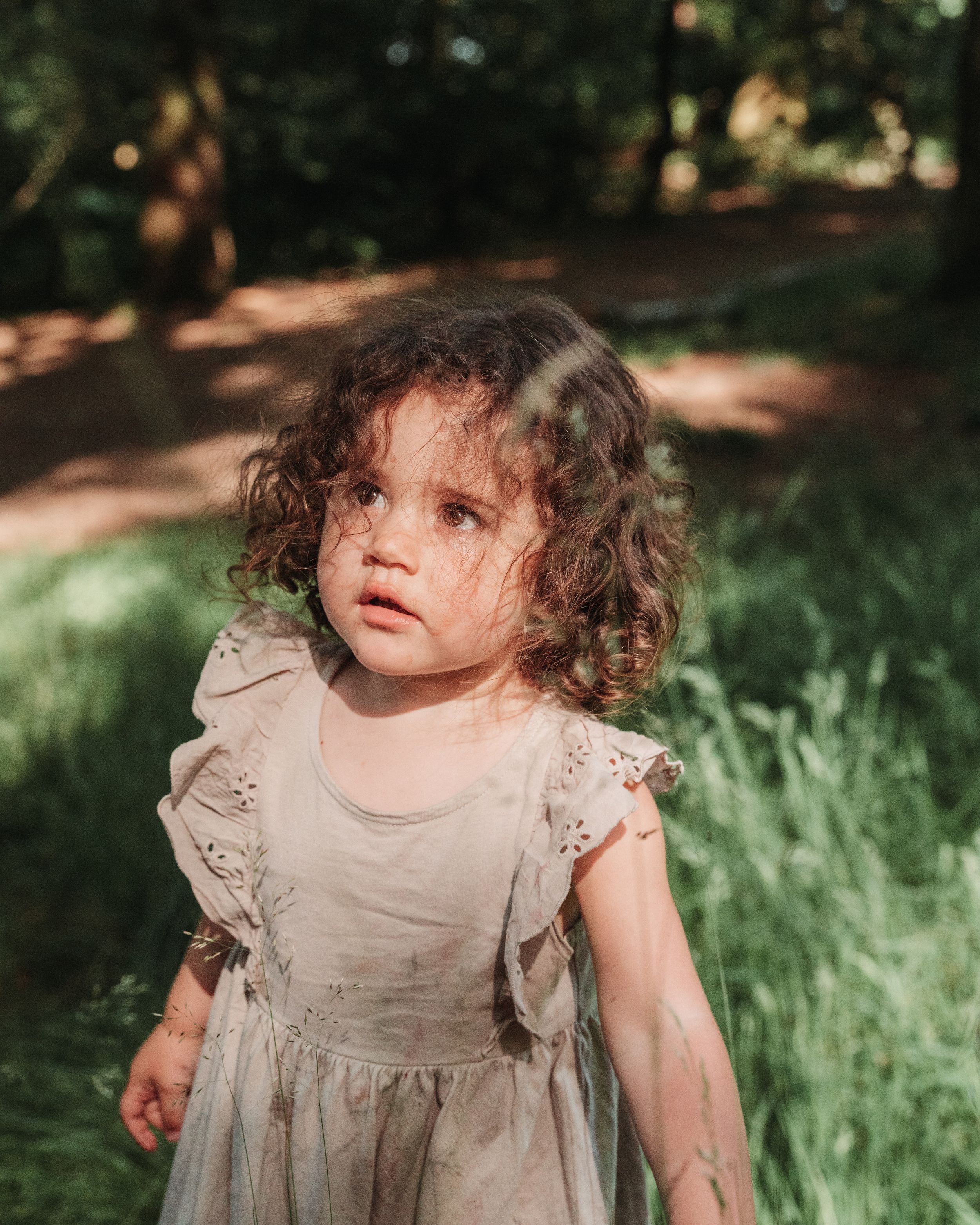 A small child wearing a ruffled beige dress stands outdoors in dappled sunlight with tall grass in the background.