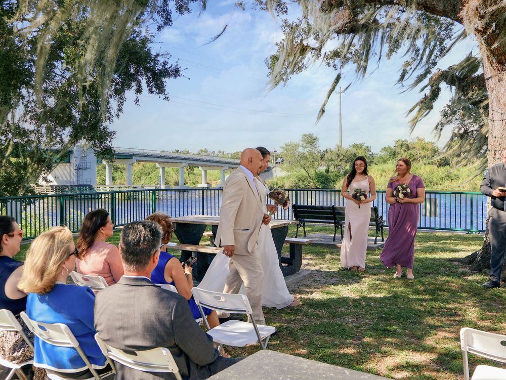 Outdoor wedding ceremony by a river, with a bride and groom walking past seated guests under shady trees.