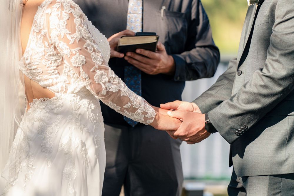 Bride and groom exchange rings during outdoor wedding ceremony while officiant holds Bible in background.