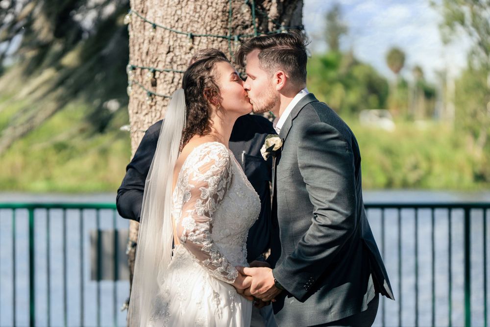 Newlyweds share first kiss in lace wedding dress and gray suit beside palm tree at outdoor wedding.