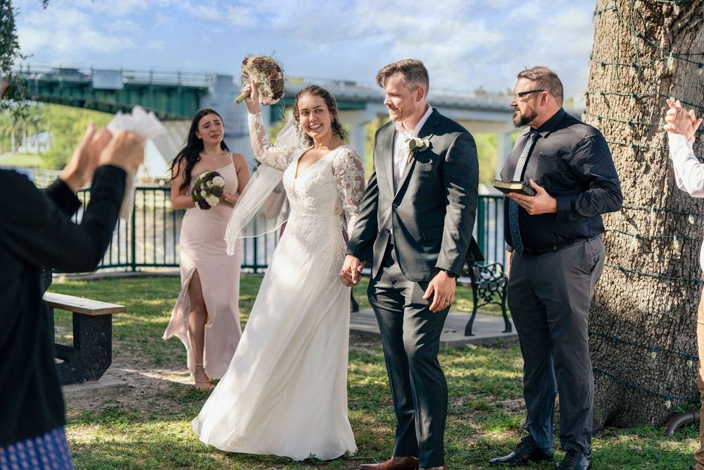 Bride and groom walk joyfully after outdoor wedding ceremony, guests celebrating with confetti, bridesmaid and officiant nearby.