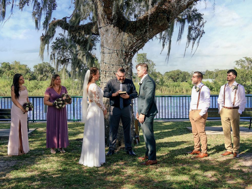 Outdoor wedding ceremony under a large oak tree with Spanish moss, bride and groom exchanging vows with wedding party.