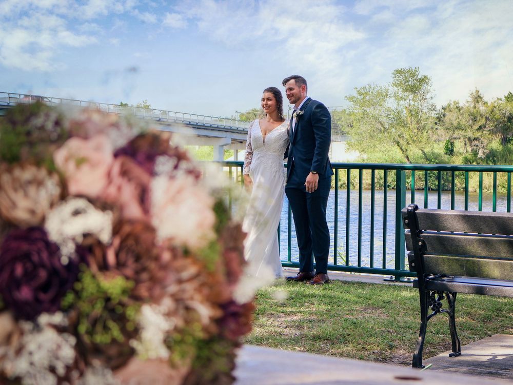 Bride and groom posing on a bridge with lush greenery, beside a floral bouquet and park bench on a sunny day.