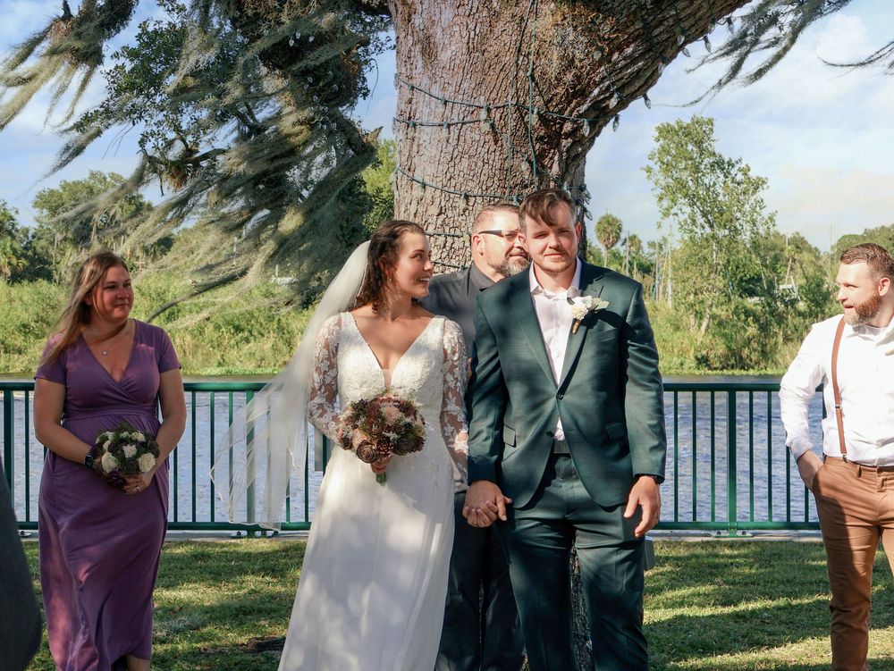 Bride and groom smiling joyfully after wedding ceremony outdoors under large tree with wedding party.