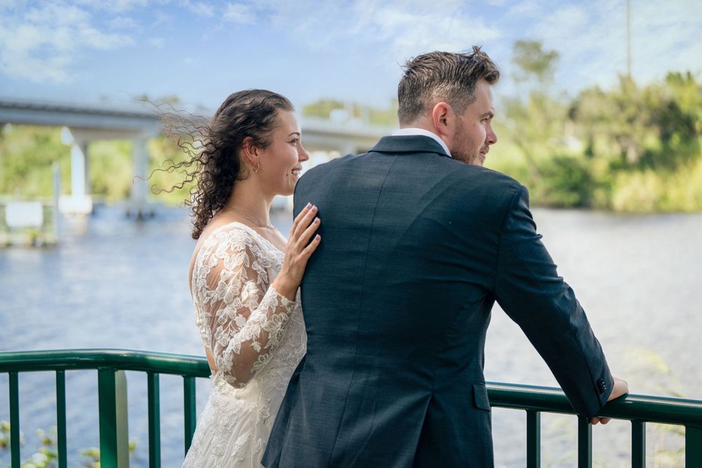 Bride in lace dress smiling at groom on waterfront bridge at outdoor wedding venue.
