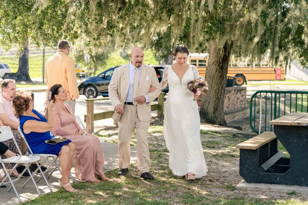 Bride walks down outdoor aisle with escort at sunny park wedding ceremony with guests seated in chairs.