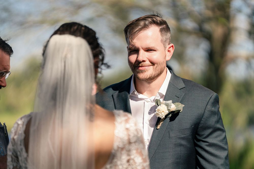 Groom smiling warmly at bride during outdoor wedding ceremony, wearing gray suit with white boutonniere.