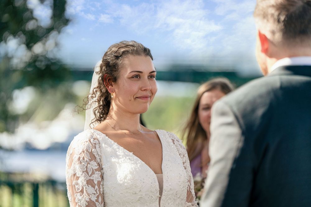 Bride in lace wedding gown smiles emotionally at groom during outdoor waterfront wedding ceremony.
