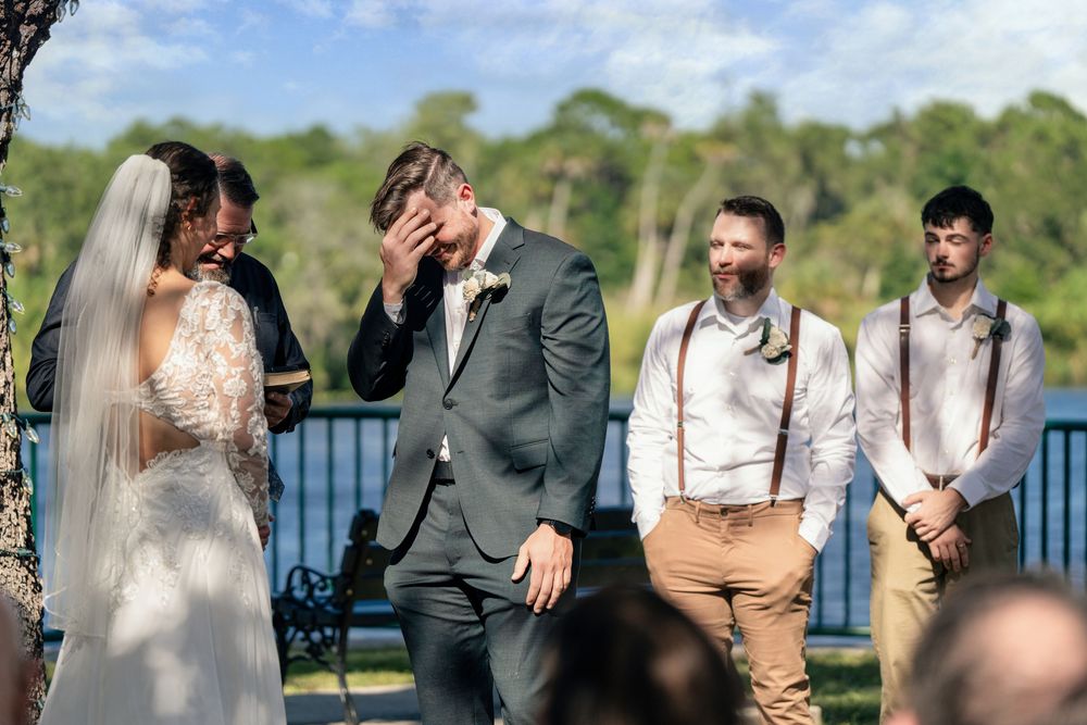 Emotional groom covering his face during outdoor wedding ceremony with bride in lace dress and groomsmen in suspenders.