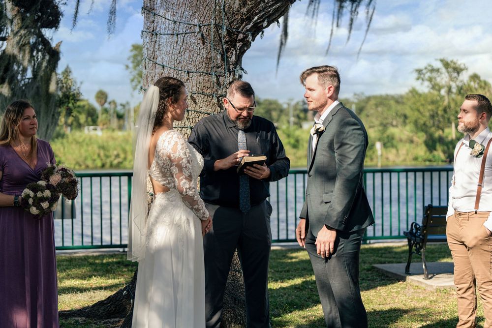 Outdoor wedding ceremony by a lake with bride in lace gown, groom in gray suit, and officiant reading vows.