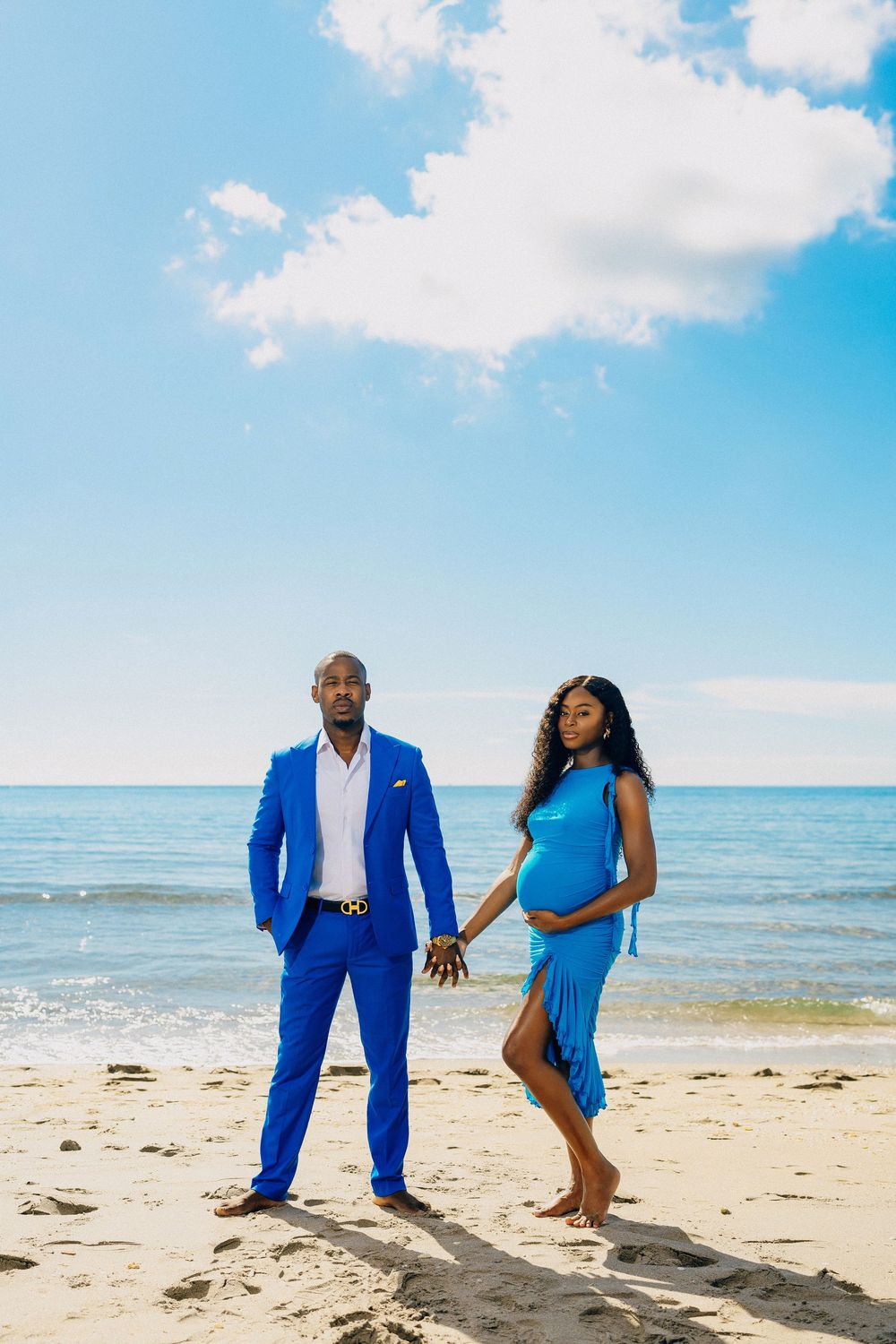 Couple in coordinating blue outfits pose together on a sunny beach with ocean and blue sky in the background.