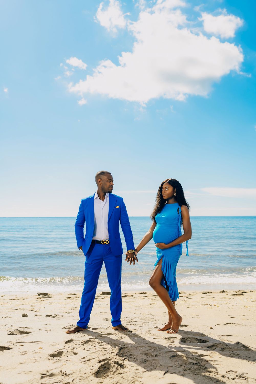A couple in coordinating blue outfits holding hands on a sunny beach with ocean and clouds in the background.