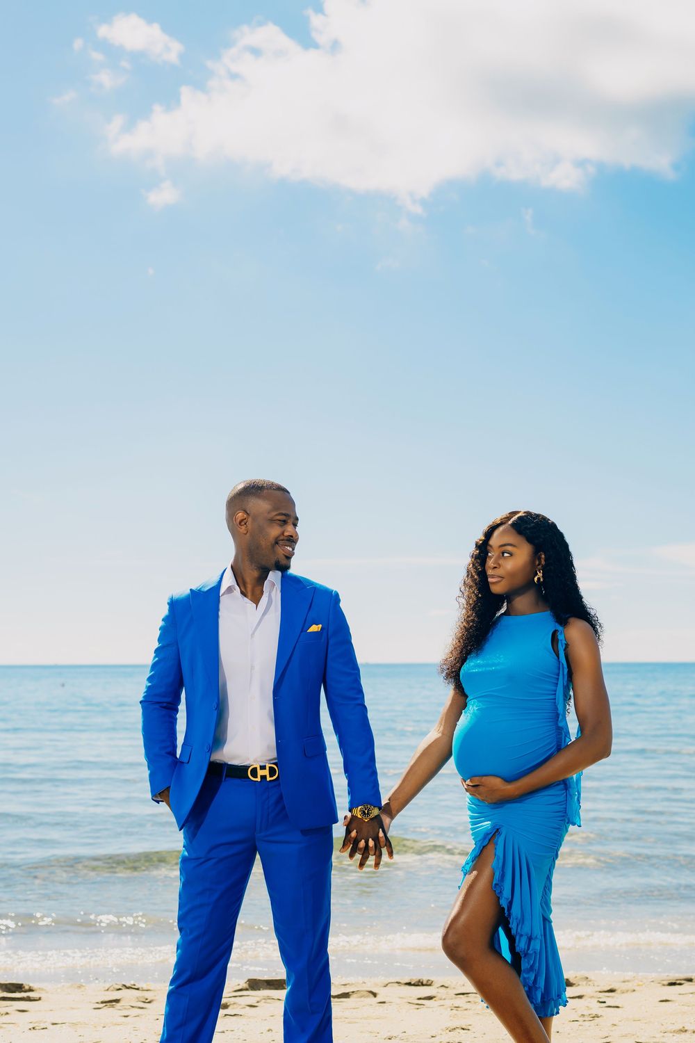 A couple holds hands on a sunny beach, with the man wearing a bright blue suit and woman in a flowing blue dress.