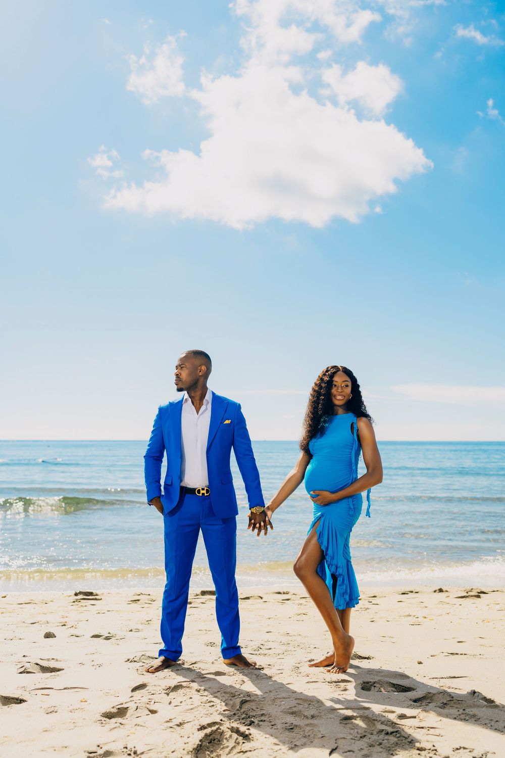 A couple holding hands on a sunny beach, with one person in a bright blue suit and the other in a flowing blue dress.