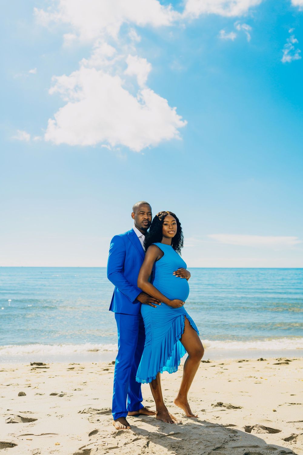 A couple in coordinating blue outfits poses for a maternity photo on a sunny beach with turquoise water and blue sky.
