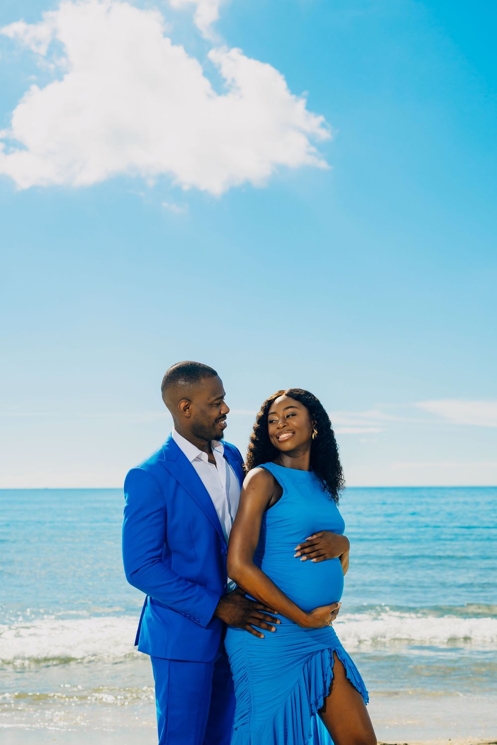 A couple in matching blue outfits shares a romantic moment on a sunny beach with ocean waves and blue sky in the background.