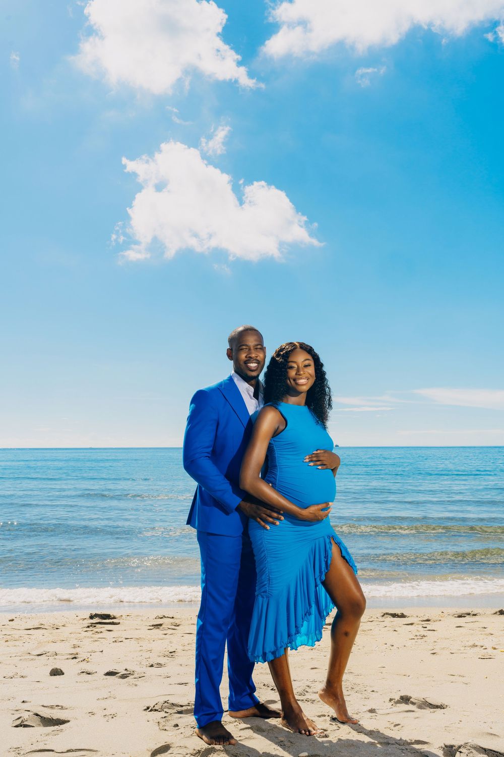 Couple in matching blue outfits embrace on a sunny beach with ocean and blue sky backdrop.