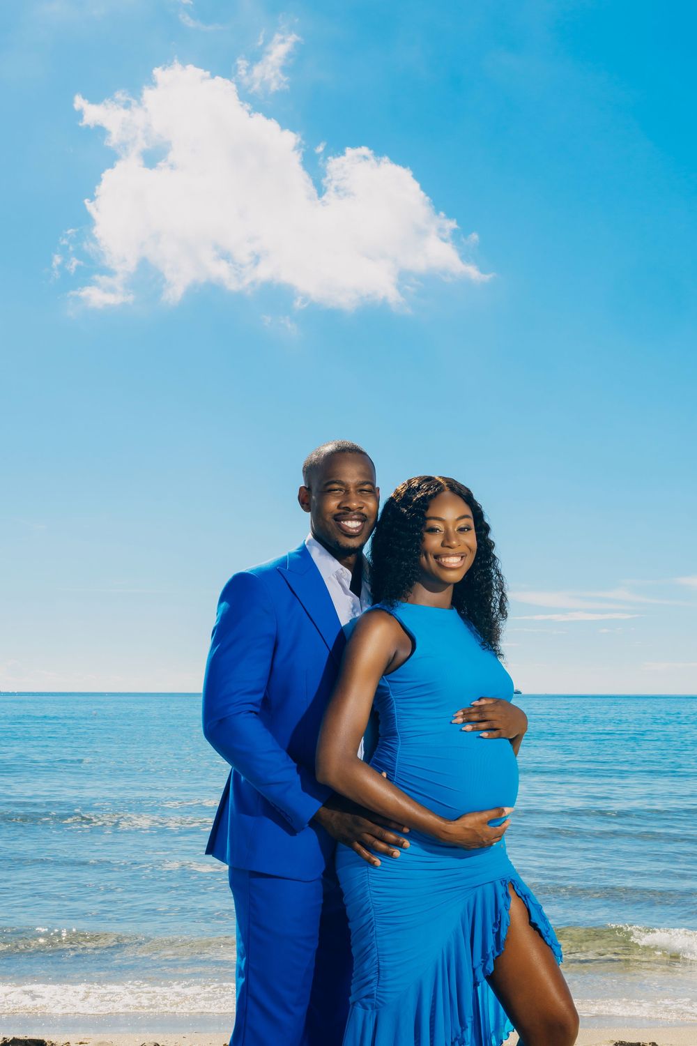 A couple in coordinating blue outfits embrace during a maternity photoshoot on a sunny beach with ocean backdrop.