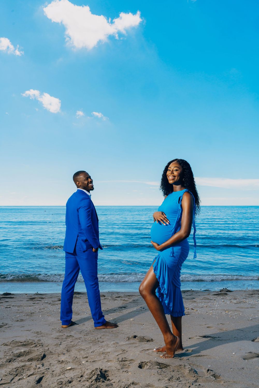 A couple in coordinating blue outfits poses for a maternity photo shoot on a sunny beach with ocean backdrop.