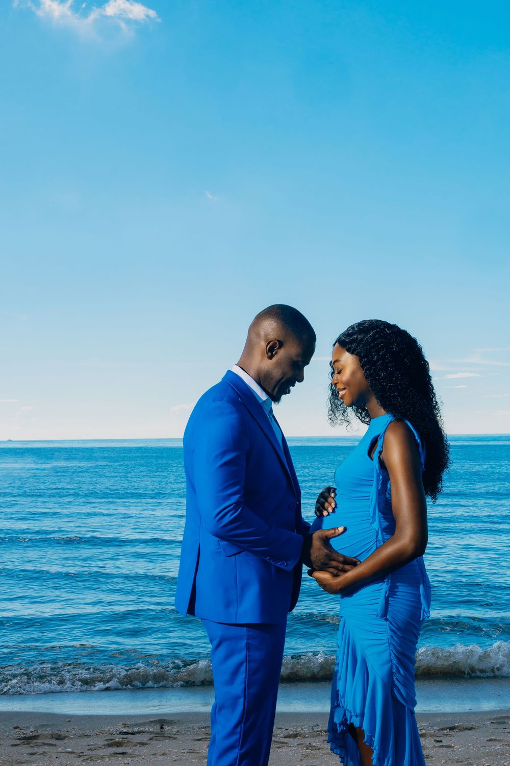 A couple in matching royal blue outfits share a romantic moment on a beach with the ocean and blue sky in the background.